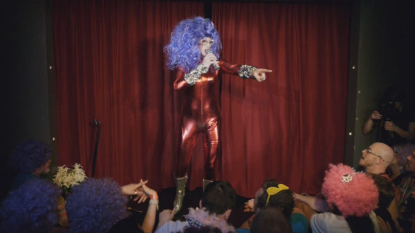 Live show video production. Dame Edna is wearing a red metallic one piece and a large purple curly wig.