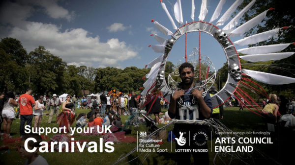 Fundraising video production. A man with a large round frame with feathers attached stands in front of people preparing for Luton carnival.