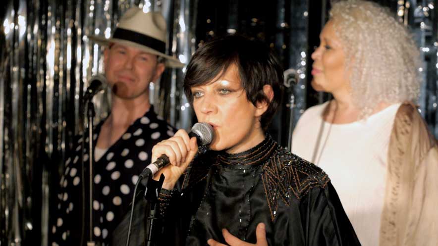 Filming live performances. Bille sings with backing singers behind her on a stage set with black and silver flitter
