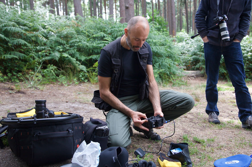 Tips for filming outdoors. Julian adjusts the settings on his Tascam DR-60 digital audio recorder in the forest surrounded by his kit. A cameraman is watching.