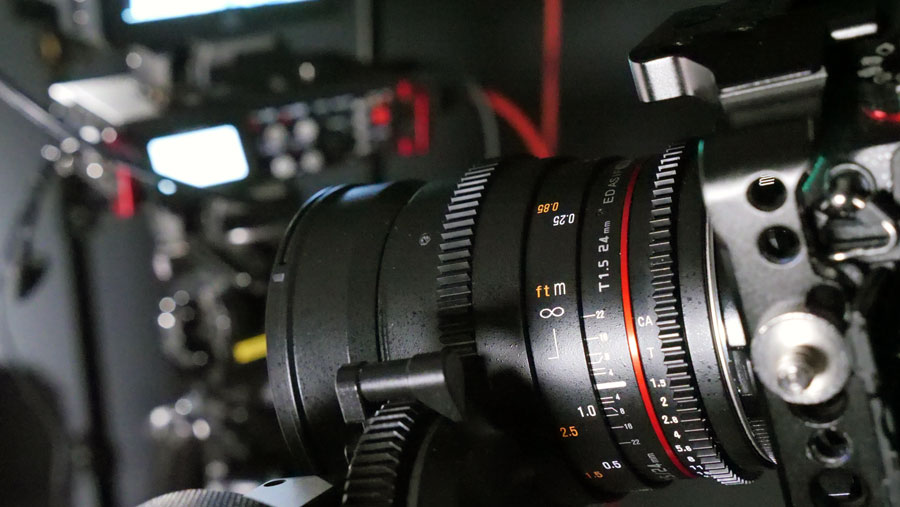 Videography filming kit list. Showing the distance and aperture graphical markings on the side of a prime lens with a focus pulling device engaged with its gear cogs. A sound recording device is out of focus in the background.