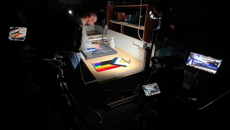 Showing Bruno Romanelli making the Pride Progress flag in fused glass, being filmed with two cameras and lit by two lights in a dark studio.