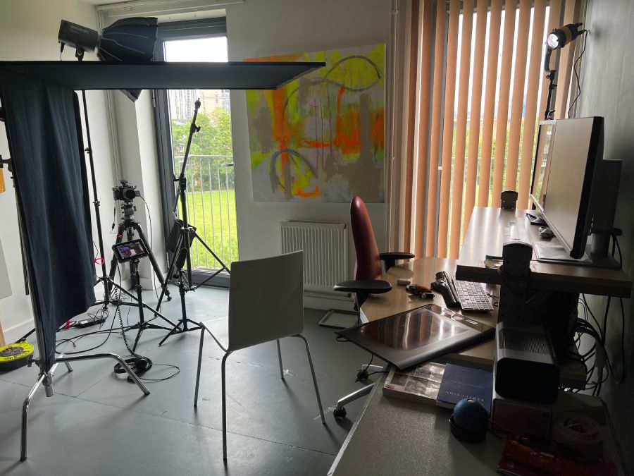 Professional talking-head video production showing a behind-the-scenes studio setup. Position of windows, stand-mounted negative-fill floppy, spot lighting, editing workstation, presenter's chair, and tripod-mounted camera.