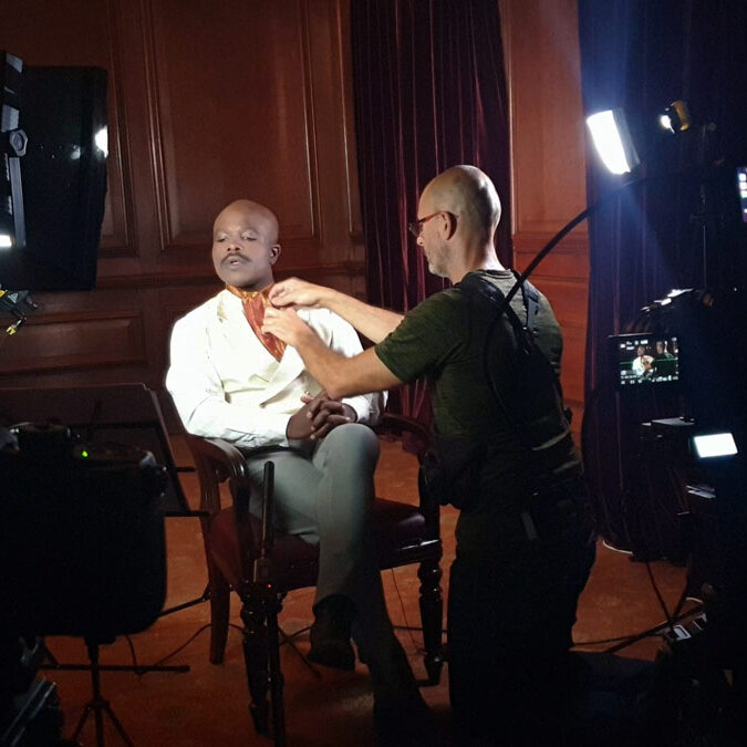 Julian-Langham-Filmmaker-adjusting-the-mic Filmmaker Julian Langham is adjusting the mic on a seated actor, showing the camera equipment and lighting.