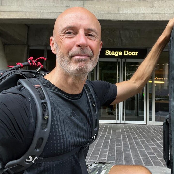Julian Langham, a videographer, is captured in a mid-shot squatting and looking at the camera while wearing a rucksack with filming equipment.